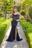 Wearing the Long Prom Dress Juliet JT2706K, a woman poses with her hands on her hips in a manicured garden. The strapless black gown features an embellished corset bodice and a high slit as she stands on a stone path.