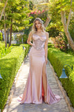 A woman wears the blush Juliet JT2706K long prom dress with a strapless embellished corset bodice and satin sheath skirt, standing on a stone garden path surrounded by lush greenery.