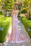 Wearing the Juliet JT2706K Long Prom Dress—a light pink, fitted satin gown with an embellished corset bodice—a woman stands on a garden stone path, glancing over her shoulder.