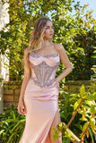 A woman stands outdoors among green plants, looking to the side, wearing the Juliet JT2706K long pink strapless prom dress with an embellished corset bodice.