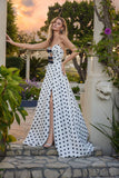 A woman in the Juliet JT2740S long prom dress in polka dot stands outdoors by a stone pillar, surrounded by greenery and flowers at sunset, wearing a strapless sweetheart neckline style with a thigh-high slit.