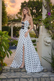 A woman wearing the Long Prom Dress: Juliet JT2740S, featuring a polka dot pattern, a-line skirt, and strapless sweetheart neckline, stands outdoors near plants and classical columns at sunset.