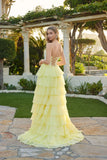 A woman stands outdoors in a garden, wearing the Juliet JT2738S long prom dress—a yellow tiered gown with a corset bodice and lace-up back—looking over her shoulder.