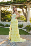 A woman stands outdoors by greenery and stone architecture, wearing the Juliet JT2738S Long Prom Dress in yellow, featuring a strapless design, tiered chiffon ruffle skirt, and high slit.