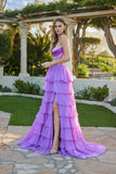 A woman stands outdoors by greenery and stone architecture, wearing the Juliet JT2738S Long Prom Dress in purple, featuring a strapless design, tiered chiffon ruffle skirt, and high slit.