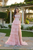 A woman stands outdoors by greenery and stone architecture, wearing the Juliet JT2738S Long Prom Dress in pink, featuring a strapless design, tiered chiffon ruffle skirt, and high slit.