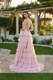 A woman in the Long Prom Dress: Juliet JT2738S, featuring a strapless corset bodice and pink chiffon ruffle skirt, stands outdoors on a stone path amid greenery and classical architecture.