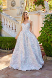 A woman stands outside among stone statues and greenery, wearing the Juliet JT2742A Long Prom Dress in Blush, featuring a fitted corset bodice, thin straps, and floral-textured fabric.