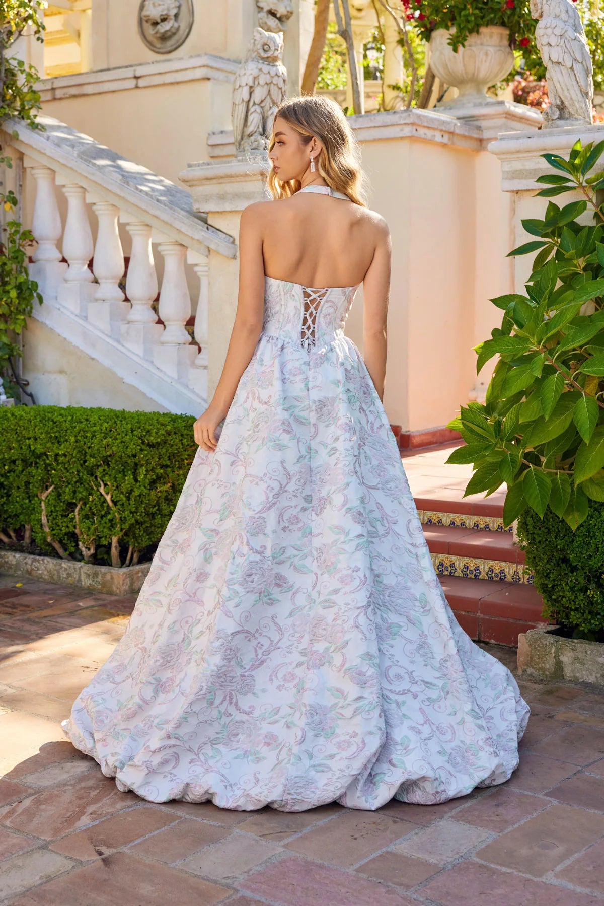 A woman in the Juliet JT2742A long prom dress stands outdoors on a tiled patio near white stairs and greenery, looking over her shoulder.