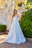 A woman wears the Juliet JT2742A Long Prom Dress, a light blue floral floor-length gown with a delicate corset bodice, standing outdoors by a stone staircase and lush greenery.