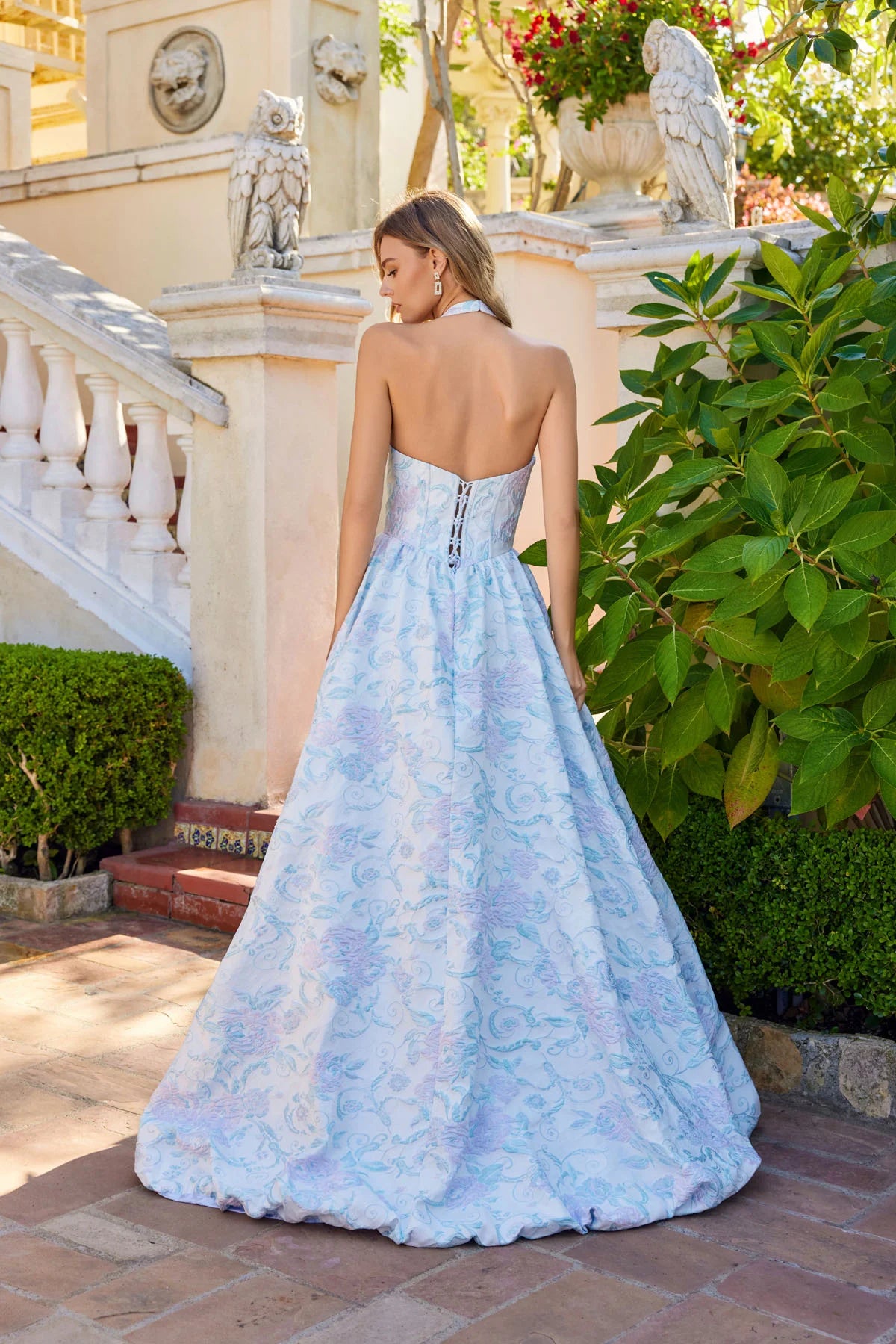 A woman wears the Juliet JT2742A Long Prom Dress, a light blue floral floor-length gown with a delicate corset bodice, standing outdoors by a stone staircase and lush greenery, looking over her shoulder.