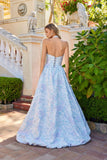 A woman wears the Juliet JT2742A Long Prom Dress, a light blue floral floor-length gown with a delicate corset bodice, standing outdoors by a stone staircase and lush greenery, looking over her shoulder.