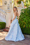 A woman wears the Juliet JT2742A Long Prom Dress, a light blue floral floor-length gown with a delicate corset bodice, standing outdoors by a stone staircase and lush greenery, looking over her shoulder.