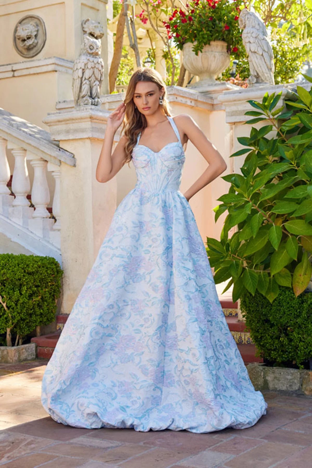 A woman stands outside among stone statues and greenery, wearing the Juliet JT2742A Long Prom Dress in Aqua Mist, featuring a fitted corset bodice, thin straps, and floral-textured fabric.