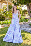 A woman stands outdoors in a garden wearing the Juliet JT2742A Long Prom Dress, featuring a strapless white design with a lavender floral pattern and an elegant corset bodice.