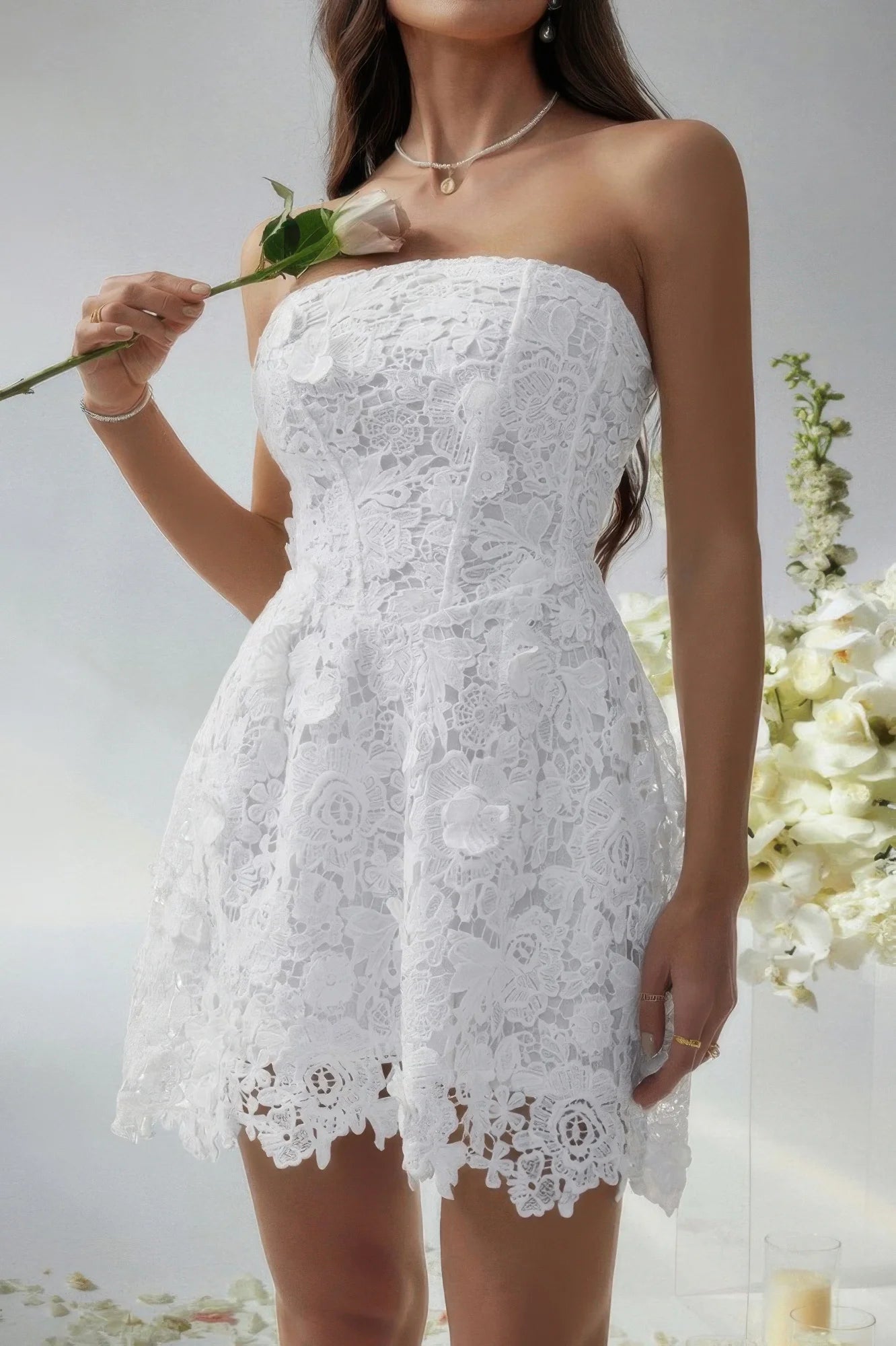 Woman wearing a white lace strapless dress holding a flower against a neutral background