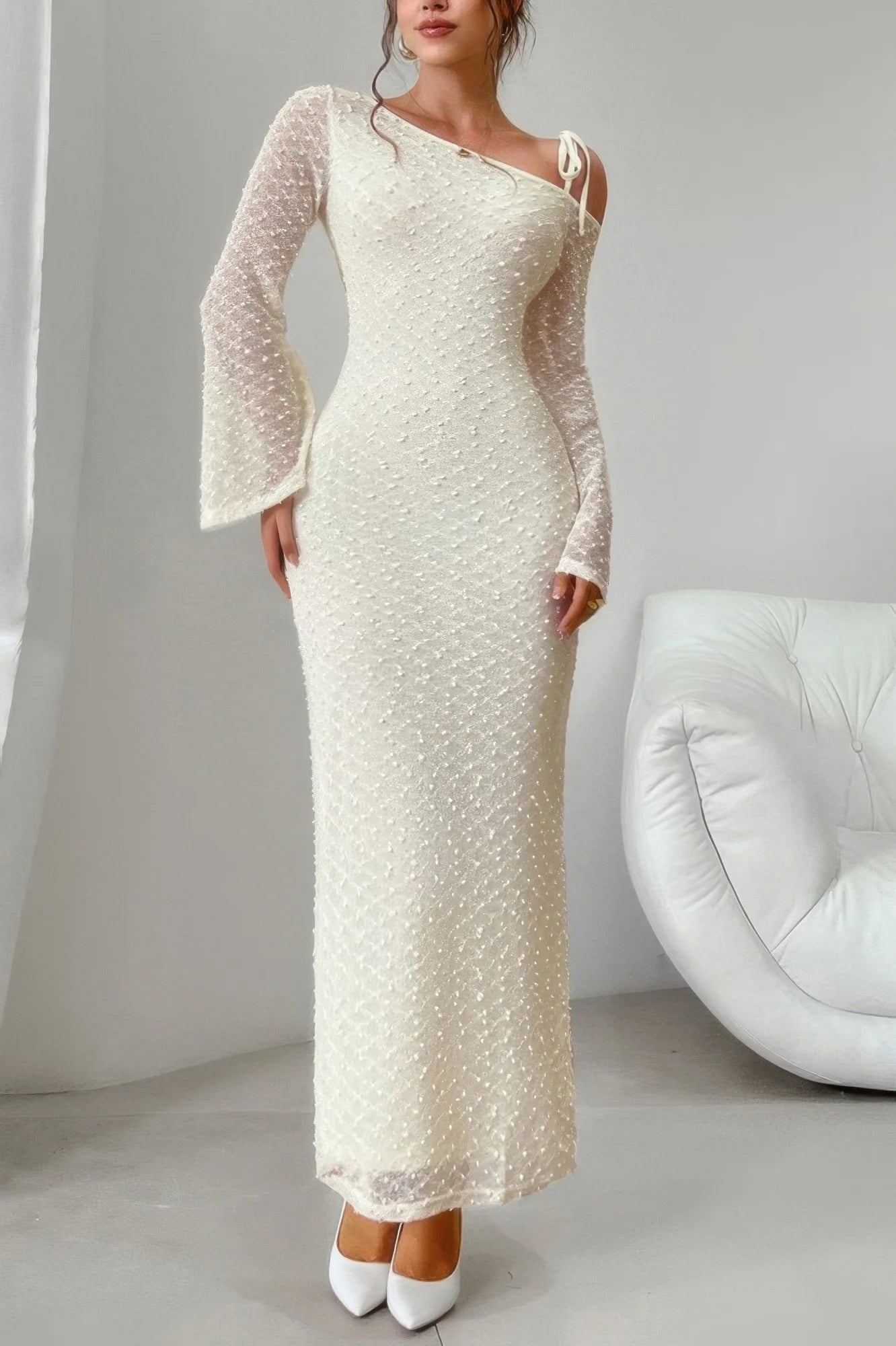 Woman wearing a long, elegant white dress with sheer sleeves in a minimalistic setting.