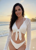 Woman in a white bikini and cover-up standing on a beach at sunset.
